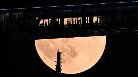 Getty Images People at the top of the Shanghai World Financial Centre in China, which resembles a bridge, as the giant Blood Moon rises above skyscrapers in the financial district. 