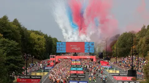North News Overhead shot of crowd of people starting race near starting line. There are overhead flare shots. Behind the starting line is a crowd of people getting ready to run. The banner around the starting line is red. 
