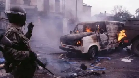 Getty A British Army soldier stands with his back turned near a burning van as debris lies around him on a Belfast street. 