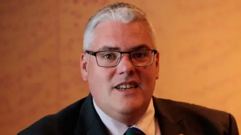 PA Media Gavin Robinson in front of an orange wall. He is wearing a suit and has grey hair.