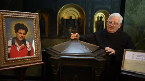A friar in a dark cloak stands next holding the lid of a font, between a framed picture of a boy in a red top and a framed baptism certificate