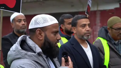 Darul Arqam Mosque and Muslim Community Center Men Taking part in a "walk of solidarity"they are holding up signs and a bearded man closest in the picture is wearing a white Muslim prayer cap and is speaking into a small microphone