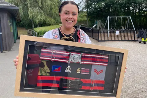 Mel is holding a large frame containing a display of her military cap badges and other items