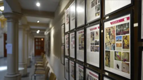 A shot of a corridor with pillars and chairs lined up, with the focus of the camera on a series of printed and framed webpages