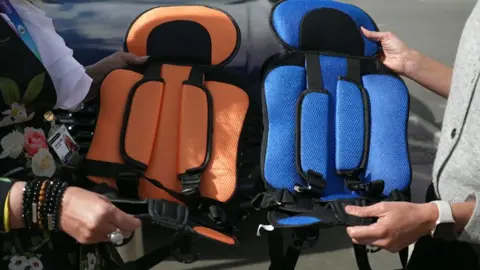 BBC Two unidentifiable people holding a child's car seat each. One on the left is orange and the other is blue. The seats are made of thin padded fabric without structure.