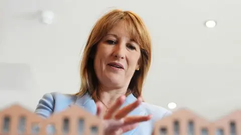 WPA Pool/Getty Images Chancellor Rachel Reeves, wearing a powder blue suit, standing with her hand over some model houses which are just seen in front of her