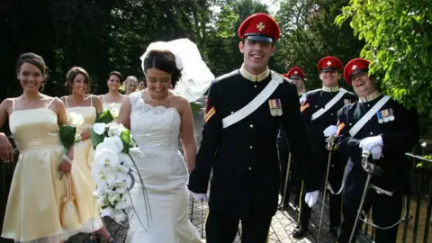 Mel Dyke Mel is wearing her wedding dress and holding hands with her husband, Chris, who is in full ceremonial military dress.