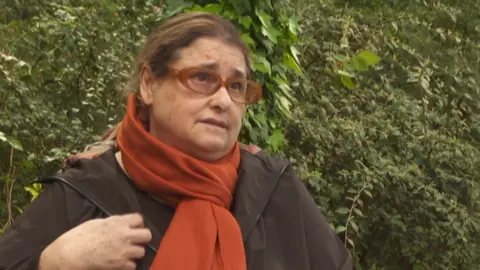 A woman in brown glasses, a brown coat and orange scarf looks to the side of the camera, stood in front of a hedge