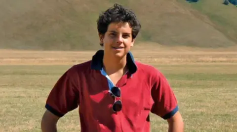 BBC A boy with dark curly hair in a red polo shit stands, smiling at the camera, with his hands on his hips, in front of a field and hills