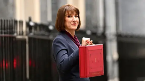 Leon Neal/Getty Images Chancellor Rachel Reeves, wearing a dark blue suit, holding the red Budget box, standing in front of the black railings on Downing Street, looking to the side and straight into the camera