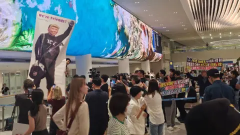 BBC/Lee-hyun Choi A large group of journalists and some protesters had gathered at Incheon International Airport ahead of their arrival. Some held up banners dismissing the raid