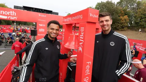 North News Jacob Murphy and Nick Pope in black tracksuits smiling as they ring a red bell with branding that says "I Bell". People in wheelchairs start racing behind them from the start line