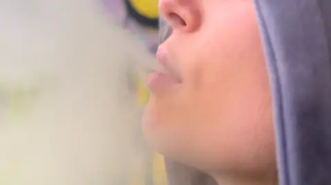 A close-up side profile of a woman vaping. She is exhaling and there is a cloud of vapour to the left of the picture. Only her nose and mouth can be seen and she has her grey jumper hood up. Yellow and purple graffiti can just be seen behind her.