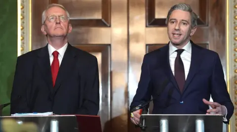 PA Media Simon Harris in a black suit jacket and red tie, he has short grey hair. A younger man, also with grey hair, is stood beside him in a navy suit, also wearing a white shirt and purple tie.
They are stood at two podiums outside a large wooden door.