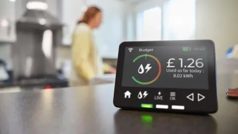 Getty Images A smart meter energy display on a kitchen surface with the screen saying £1.26 of energy has been used today. A woman in the kitchen is blurred in the background.