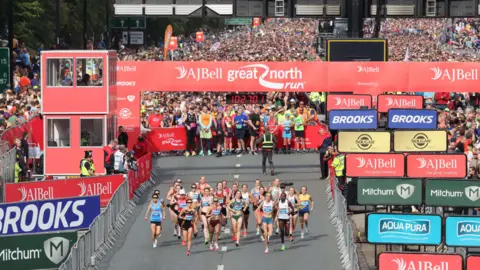 North News Overhead shot of elite women running near the starting line. They are all in running gear looking focused. Behind the starting line is a crowd of people getting ready to run. The banner around the starting line is red. 