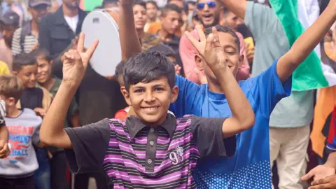 Getty Images Palestinian children celebrate in Al-Mawasi, Rafah, Gaza Strip, on October 9, 2025, following news of a new Gaza ceasefire deal. A boy at the front of the shot is wearing a black and purple striped top. He is smiling at the camera and making the 'peace' sign with his fingers. 