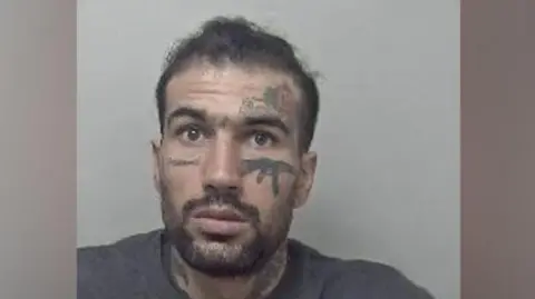Metropolitan Police Custody photo of Fayaz Khan, who has short dark hair, a beard and moustache.  He has tattoos on his face, including one of an AK-47 gun beneath his left eye. He is wearing a grey jumper.
