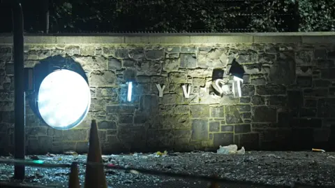 PA Media A stone wall illuminated by a bright light - a sign that was on the wall hangs from it