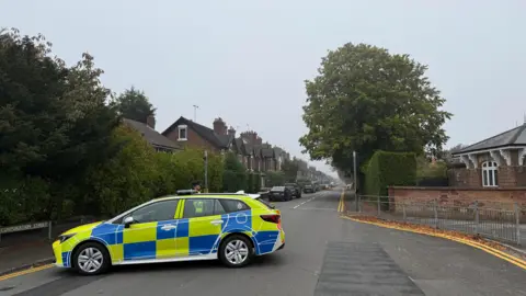 A yellow and blue Staffordshire Police car is parked across Corporation Street in Staffordshire. An officer can be seen standing next to the vehicle