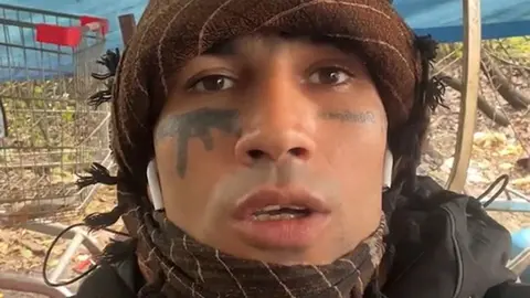 PA Media Fayaz Khan looking at the camera. He has a brown bandana wrapped around his head and throat. He is wearing small white earphones. He is speaking in a selfie-style video and has tattoos under his eyes, one of which depicts a gun.