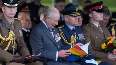 PA Media The king sits in the front row of the dedication ceremony flanked by uniformed members of the armed forces. He is looking down reading an open book of service with the LGBT rainbow on the front.