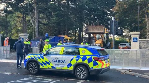A Garda car parked on a road with gardaí and men in hi-vis clothing standing behind it. There is a row of trees in the background. 