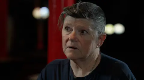 A head and shoulders shot of Pádraigín, who is looking off to the side of the camera. She has short graying hair brushed back at the front and is wearing a dark T-shirt.