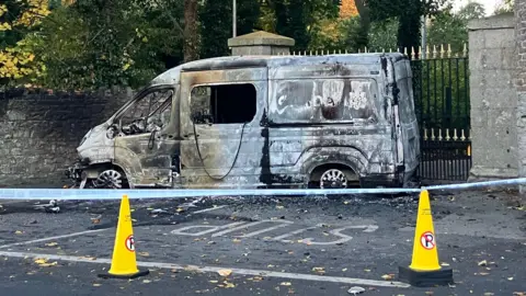 A burnt out police vehicle behind a cordon