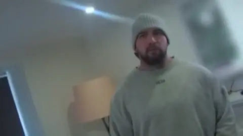Gwent Police Bodycam footage of a man in a grey sweather and grey beanie hat stands in his living room with a lamp and a television behind him