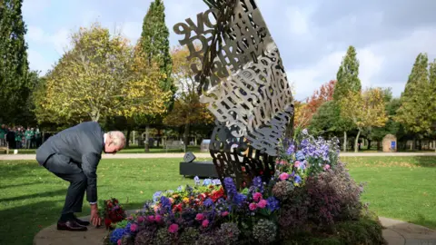 Reuters The King stoops to lay flowers at the foot of a twisted metal sculpture made up of lots of individual words and phrases, shaped like a large scrunched piece of paper. It is a sunny day at the arboretum and the trees are bathed in sunlight in the background.
