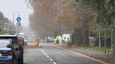 BBC Corporation Street in Stafford, with trees lining each side of the road. There are cars lining the left hand side of the road, with emergency service vehicles and workers visible on the left hand side. 