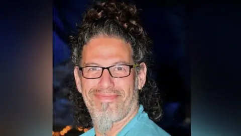 Hostages and Missing Families Forum Image shows Ronen Engel in a turquoise blue shirt, with a beard and black glasses smiling with a topknot of dark brown curls on top of his head