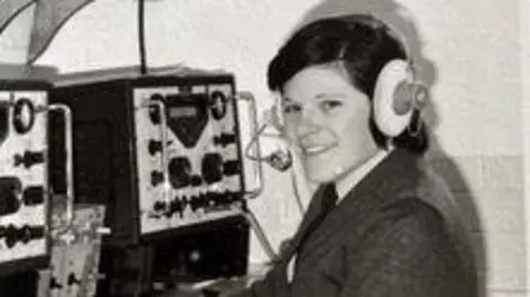 Pádraigín Ní Rághillíg A grainy black-and-white archive photo shows a young Pádraigín smiling at the camera behind a large machine on a desk covered in dials and switches. She is wearing a suit and tie and has a large headset on her head. 