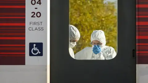 Getty Images Members of a forensics team, wearing white suits and blue latex gloves, are seen in the reflection of a train window at Huntingdon station on Sunday