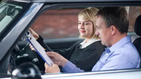 Getty Images A young woman taking a driving test. She has blonde hair and is wearing a dark grey jumper. Her tester is a man in a check blue shirt. He is showing her a document on a clipboard.