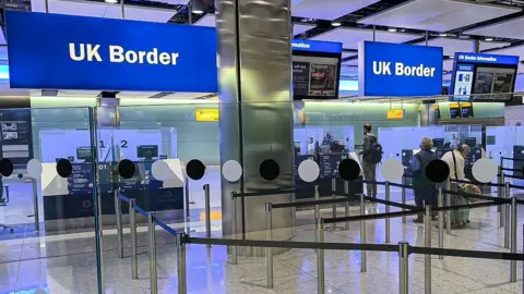 NurPhoto via Getty Images Uk Border at Heathrow airport. two blue signs that read UK border. Below it are the passport control desks