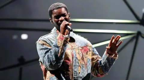 Getty Images Dave is singing in to a microphone while performing on stage. He is wearing a distressed denim jacket. 