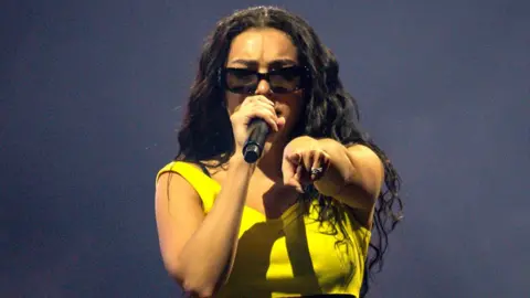 Getty Images Charli XCX wears a yellow strappy top. She has long brown wavy hair and is wearing black sunglasses. She is singing in to a microphone and pointing at the camera. 
