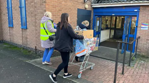 One person pushing a trolley full of bags and boxes of food. Two other people can be seen behind. They are walking towards a blue doorway.