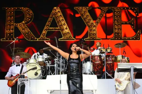 Getty Images Raye wears a black strappy dress. She is performing on stage and singing in to a microphone. There is a band playing behind her and there is a signing saying "Raye" behind her. 
