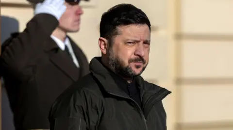 Reuters Zelensky, who has short black hair and short black facial hair, wears a black coat in front of a building with beige walls. Behind him a man in uniform stands to attentoin.
