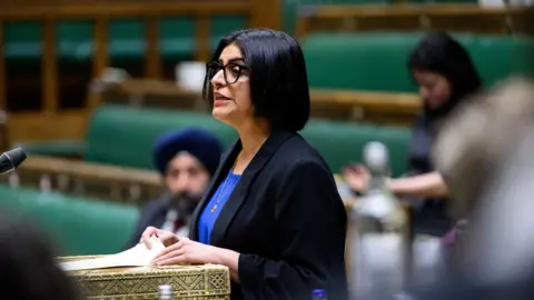 House of Commons Home Secretary Shabana Mahmood in the Commons. She has shoulder-length black hair and is wearing a black suit jacket and blue shirt. A few other MPs are visible in the blurred background.