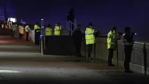 BBC/David Rimmington A row of police officers standing by a sea wall shining lights out to sea. 